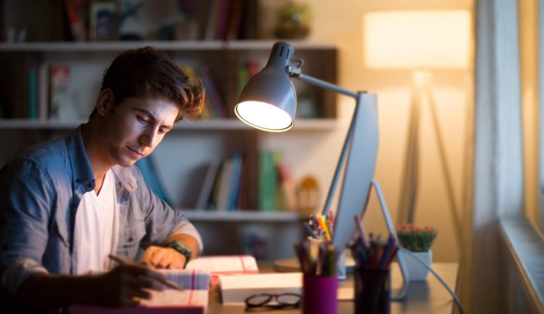 Um homem estudando em uma mesa com um computador, lapis, livros e uma luminária