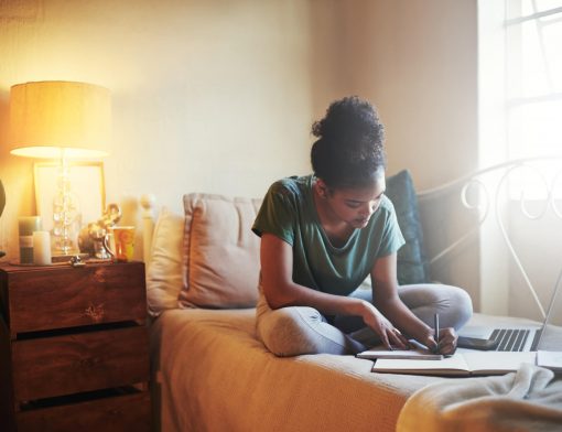 Uma menina sentada na cama anotando algo em um caderno e um notebook em cima da cama