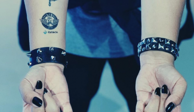 Duas mãos com unhas pretas, pulseiras punk e tatuagens fazendo um sinal de "rock"
