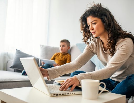 Uma mulher trabalhando no regime home office com uma criança sentada no sofá ao fundo