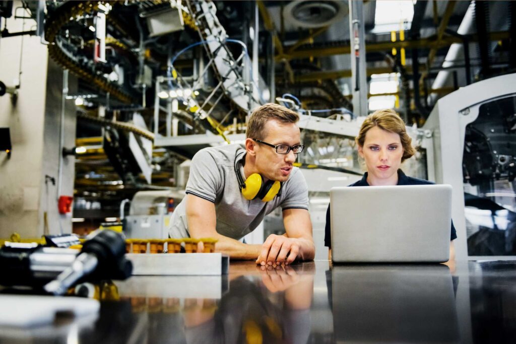imagem de uma indústria com equipamentos ao fundo e no primeiro plano um homem e uma mulher conversando olhando um notebook
