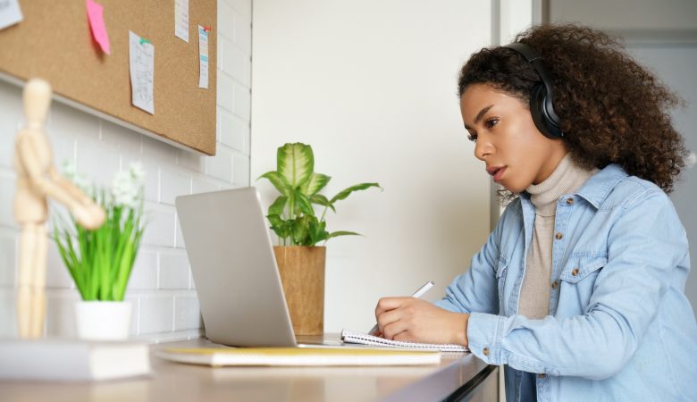Uma menina com um fone de ouvido assistindo algo pela tela do computador e fazendo anotações em um caderno