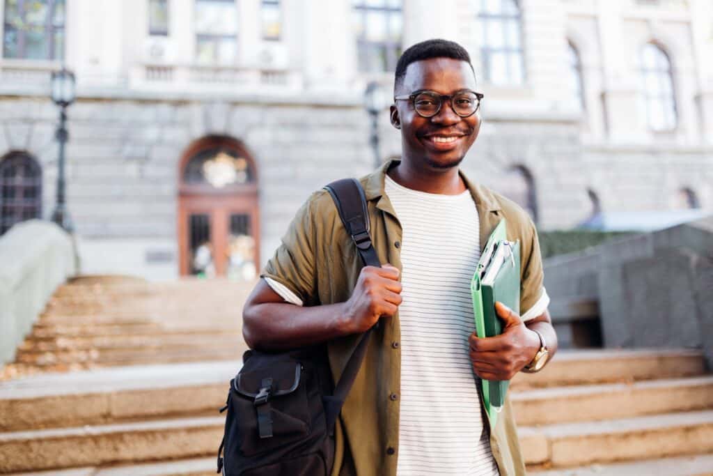 homem jovem preto está sorrindo, segura livros e mochila com um prédio ao fundo