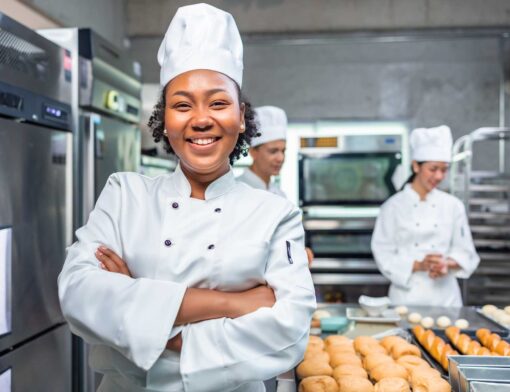 mulher preta está vestida como chef e tem os braços cruzados no primeiro plano; ao fundo é possível ver dois outros cozinheiros, equipamentos de cozinha e pães na mesa