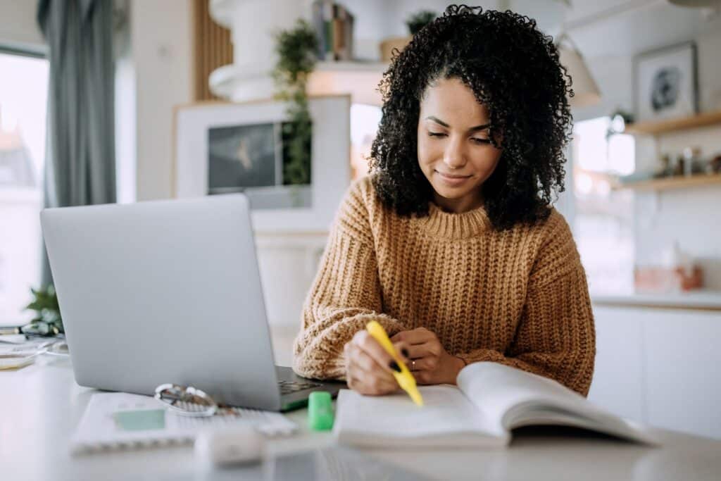 mulher negra usa blusa de tricô marrom claro, está grifando um livro aberto; ao lado tem um notebook cinza, papeis e óculos na mesa e ao fundo há cenário de um apartamento.