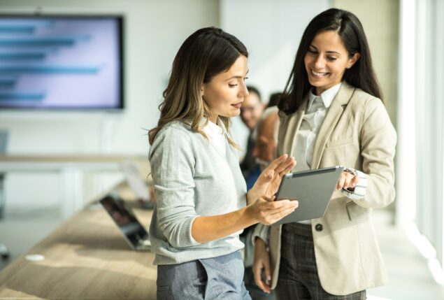 Jovens mulheres conversam em ambiente corporativo enquanto olham para um tablet na mão de uma delas.