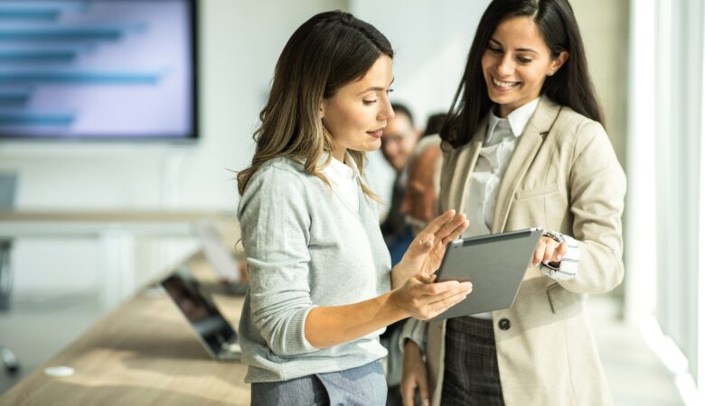 Jovens mulheres conversam em ambiente corporativo enquanto olham para um tablet na mão de uma delas.