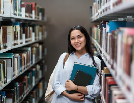 Garota jovem sorridente segura livros em biblioteca.