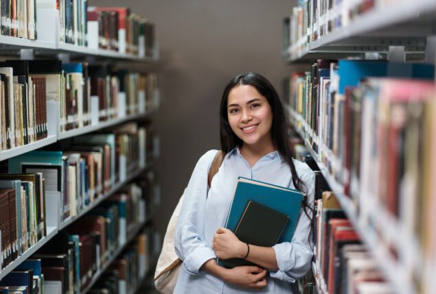 Garota jovem sorridente segura livros em biblioteca.