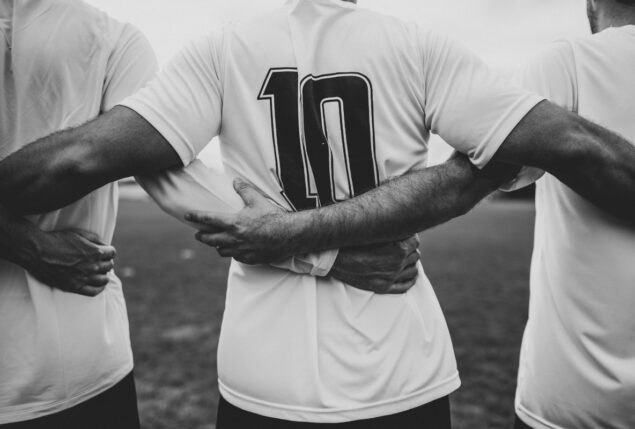 Foto em preto e branca de jogadores de futebol de costas em abraço em grupo